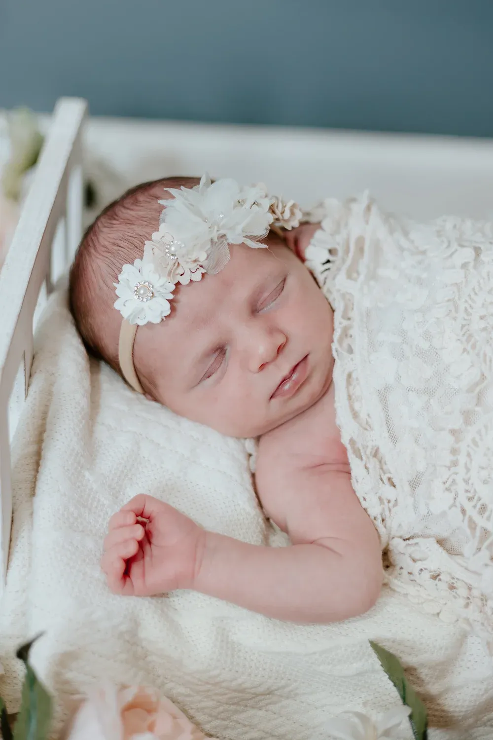 Newborn sleeping in a lace wrap with a floral headband in a wooden cradle